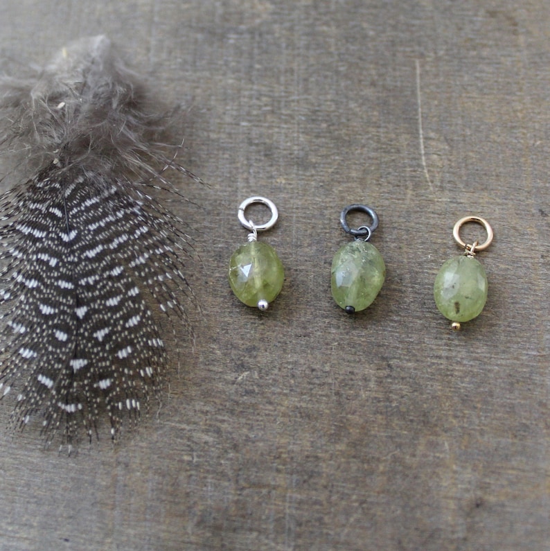 May include: Three oval-shaped green gemstone charms with different colored metal jump rings. The charms are on a wooden surface.