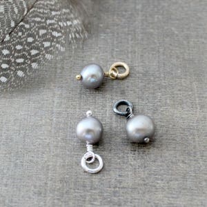 May include: Three gray pearl charms with different metal findings. The charms are on a gray surface.