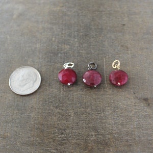 May include: Three round red gemstone charms with different metal settings. The charms are on a brown wooden surface.