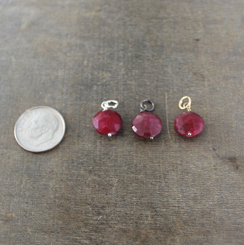 May include: Three round red gemstone charms with different metal settings. The charms are on a brown wooden surface.