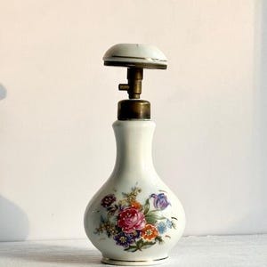 May include: A white ceramic perfume bottle with a floral design featuring red, pink, and purple roses. The bottle has a brass-colored top and a rounded shape, standing approximately 6 inches tall.