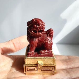 May include: A small, intricately carved red sculpture of a mythical lion-like creature, atop a gold-colored rectangular box. The sculpture is detailed with sharp features and sits on a box with a geometric design.