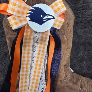 May include: A decorative boot accessory featuring a white circle with a navy blue raven graphic. The accessory includes orange and white gingham ribbon, orange and navy blue ribbons, and white lace, attached to a brown leather boot.