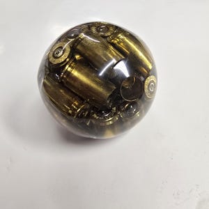 May include: A clear, spherical resin paperweight encasing numerous brass bullet casings. The casings are arranged within the transparent sphere, creating a unique and industrial aesthetic. The brass color contrasts with the clear resin.