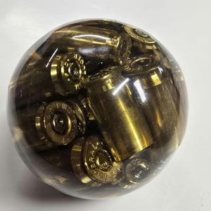 May include: A clear, spherical paperweight encasing numerous brass bullet casings. The casings are arranged throughout the sphere, creating a unique and industrial aesthetic. The brass color of the casings contrasts with the clear resin.