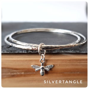 May include: Two silver hammered bangle bracelets with a silver bee charm hanging from the bottom. The bracelets are stacked on top of each other and are on a wooden surface. The text "SILVERTANGLE" is visible at the bottom of the image.