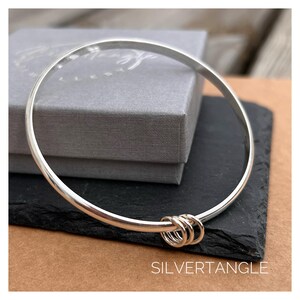 May include: A silver bangle with three small linked rings. The bracelet sits on a black surface, with a grey box behind it. The word "SILVERTANGLE" is printed in white at the bottom of the image.