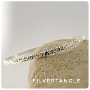 May include: A silver bangle bracelet with a textured, hammered finish. The bracelet is resting on a light brown rock.