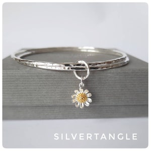 May include: A silver bangle bracelet with a hammered finish and a delicate daisy charm. The charm is made of silver and gold and has a yellow centre.