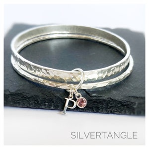 May include: Two silver hammered bangle bracelets with a silver charm featuring the letter "P" and a pink crystal. The charm is hanging from the bracelets. The bracelets are on a black surface. The text "SILVERTANGLE" is at the bottom of the image.
