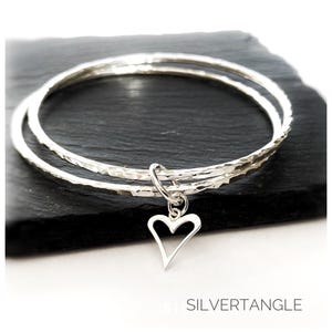 May include: Two silver bangle bracelets with a hammered finish and a small open heart charm hanging from the bottom.