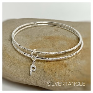 May include: A silver bangle bracelet with a hammered finish. The bracelet is double-layered and has a small charm with the letter "P" set with clear stones.