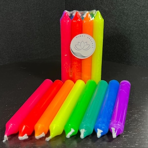 May include: A collection of colorful, rainbow-themed candles. The candles are arranged in a gradient of colors, from pink to purple. The candles are packaged together and also displayed individually. The packaging has a white label with the text "Kinky Essence".