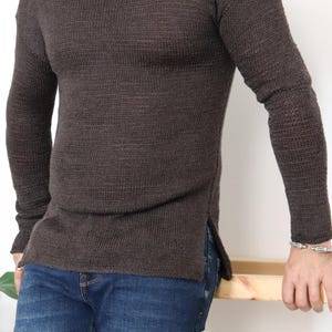 以下が含まれることがあります： リブ編みでサイドスリットが入ったブラウンのロングスリーブセーター。ブルーのジーンズの上に着ています。