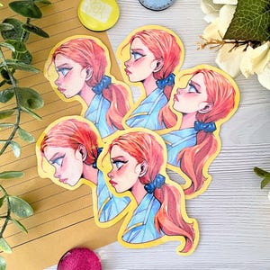May include: A collection of stickers featuring a stylized illustration of a young woman with red hair in a ponytail, wearing a blue top and a blue bow. The stickers have a yellow border and are arranged on a light-colored surface.