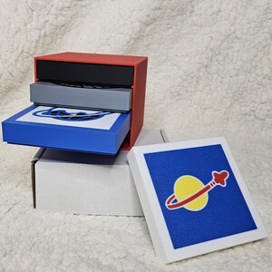 May include: A red, gray, and blue layered box with a white box with a blue background and a yellow and red planet design on top.