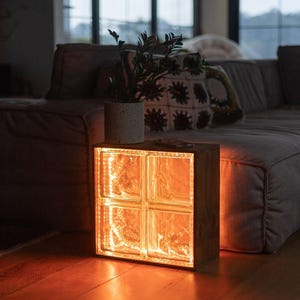 Può includere: Una lampada con cornice in legno con quattro blocchi di vetro, illuminata da un caldo bagliore arancione. Una piccola pianta in vaso è appoggiata sopra la lampada, accanto a un telefono. La lampada è su un pavimento in legno, con un divano grigio sullo sfondo.