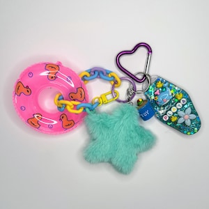 Puede incluir: Un flotador de piscina inflable rosa con patos amarillos, una estrella peluda turquesa, una cadena azul y amarilla, un llavero en forma de corazón morado y un llavero azul y verde con la palabra "PLAYYA" y una flor blanca.