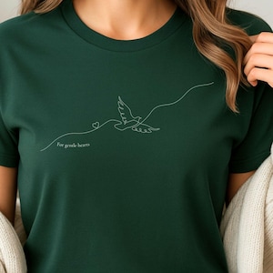 Könnte beinhalten: Dunkelgrünes T-Shirt mit einer weißen Strichzeichnung einer fliegenden Taube, einem Herz und dem Text "For gentle hearts". Das Design ist minimalistisch und elegant, geeignet für Freizeitkleidung.