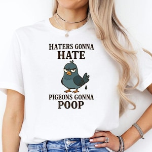 Könnte beinhalten: Ein weißes kurzärmeliges T-Shirt mit einer Grafik eines mürrischen grauen Taubenvogels mit orangefarbenen Füßen. Der Text über dem Vogel lautet HATERS GONNA HATE und der Text darunter lautet PIGEONS GONNA POOP in dunkelbrauner Schrift.