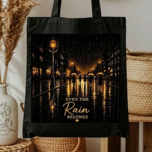 Könnte beinhalten: Eine schwarze Tragetasche mit einem Grafikdruck einer regnerischen Stadtstraße bei Nacht, leuchtenden Straßenlaternen und Silhouetten von Menschen mit Regenschirmen. Der Text lautet EVEN THE Rain BELONGS mit einem kleinen Herzsymbol.