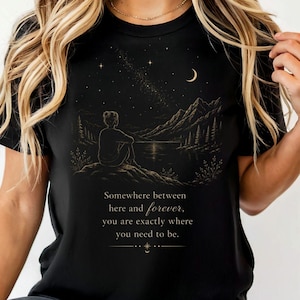 Könnte beinhalten: Ein schwarzes T-Shirt mit einer goldfarbenen Grafik, die eine Person zeigt, die auf einem Felsen sitzt und auf einen See und Berge unter einem Sternenhimmel mit Mondsichel blickt. Der Text lautet: Somewhere between here and forever, you are exactly where you need to be.