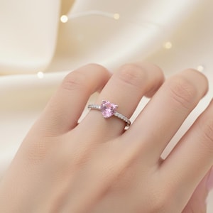 Könnte beinhalten: Ein silberner Ring mit einem herzförmigen rosa Edelstein. Das Band ist mit kleinen, klaren Steinen besetzt. Der Ring wird auf einer Hand vor einem weichen, neutralen Hintergrund gezeigt.