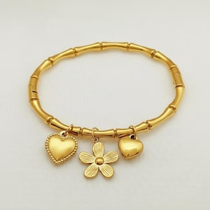 May include: Gold-colored bracelet with a bamboo-style design. The bracelet features three charms: two hearts and a flower. The bracelet is made of a shiny, metallic material and is displayed on a white background.