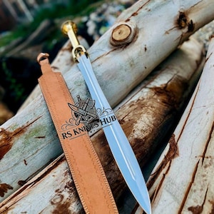 May include: A silver sword with a gold handle and a brown leather sheath. The sword is resting on a pile of wooden logs. The sheath is stitched along the edges. The text "RS KNIVES HUB" is visible on the sheath.