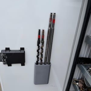May include: A collection of drill bits of varying sizes and styles, including spiral and straight designs. The bits are held in a gray holder and feature red and black accents. The image is taken inside a white cabinet.
