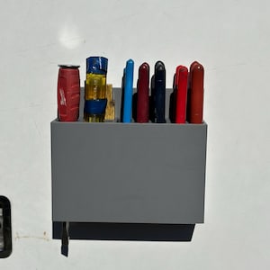 Könnte beinhalten: Ein grauer, wandmontierter Werkzeugorganizer mit einer Vielzahl von Werkzeugen, darunter ein roter Schraubendreher, eine gelbe Wasserwaage und mehrere blaue, rote und schwarze Schraubendreher.