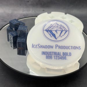 May include: A white circular silicone mold with the blue text IceShadow Productions, Industrial Bold, 8D6 123456, and a mountain graphic. Next to it are several small, dark blue cube-shaped dice with numbers engraved on their sides.