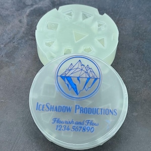 Op de afbeelding: Een doorzichtige plastic container met een blauw-wit logo met de tekst "IceShadow Productions, Flourish and Flow, 1234 567890". De container heeft een deksel met een mal om ijsblokjes te maken.