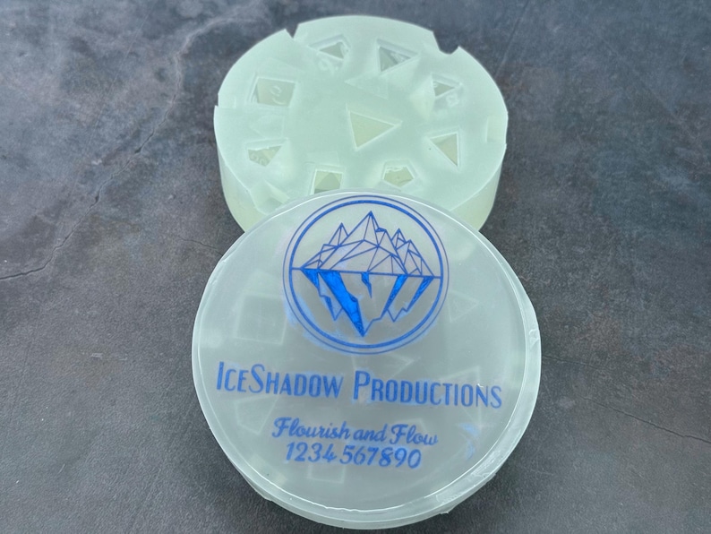 Op de afbeelding: Een doorzichtige plastic container met een blauw-wit logo met de tekst "IceShadow Productions, Flourish and Flow, 1234 567890". De container heeft een deksel met een mal om ijsblokjes te maken.