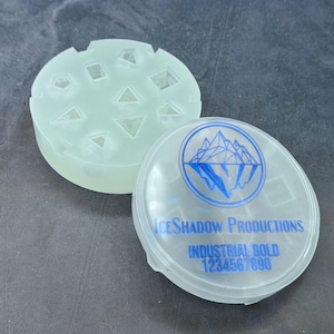 Op de afbeelding: Een doorzichtige plastic container met een blauw logo waarop "IceShadow Productions Industrial Bold 1234567890" staat. De container heeft een deksel met een mal voor zeven dobbelstenen.