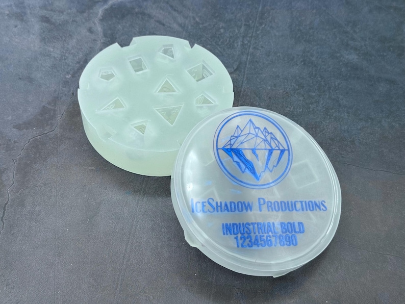 Op de afbeelding: Een doorzichtige plastic container met een blauw logo waarop "IceShadow Productions Industrial Bold 1234567890" staat. De container heeft een deksel met een mal voor zeven dobbelstenen.