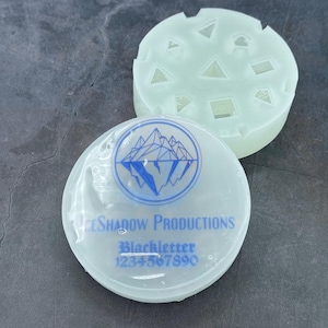 Op de afbeelding: Twee witte siliconen mallen. E&eacute;n mal heeft een bergketenontwerp met de tekst "Ice Shadow Productions" en "Blackletter 1234567890". De andere mal heeft een verscheidenheid aan geometrische vormen.