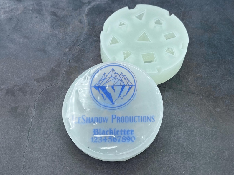 Op de afbeelding: Twee witte siliconen mallen. E&eacute;n mal heeft een bergketenontwerp met de tekst "Ice Shadow Productions" en "Blackletter 1234567890". De andere mal heeft een verscheidenheid aan geometrische vormen.