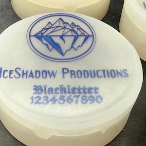 Op de afbeelding: Ronde witte container met een blauw logo van bergen en de tekst "ICE SHADOW PRODUCTIONS" en "Blackletter 1234567890" gedrukt op de bovenkant.