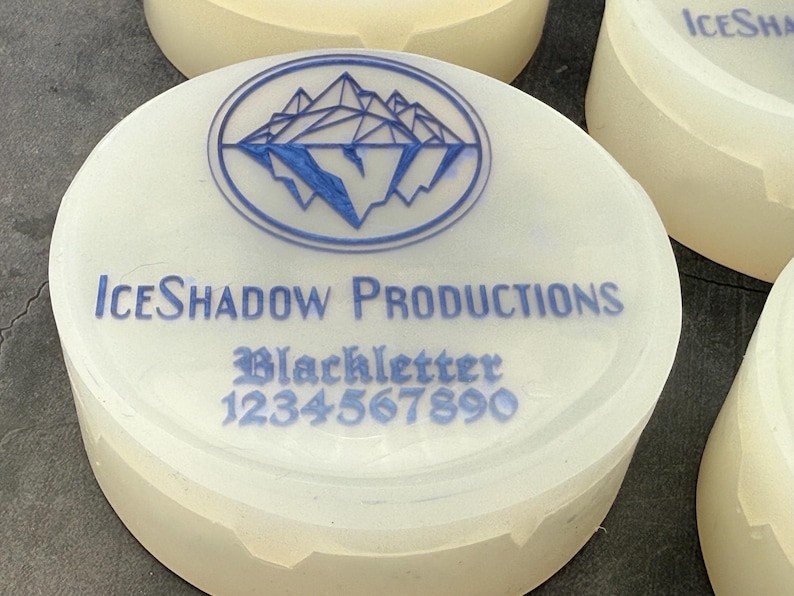 Op de afbeelding: Ronde witte container met een blauw logo van bergen en de tekst "ICE SHADOW PRODUCTIONS" en "Blackletter 1234567890" gedrukt op de bovenkant.
