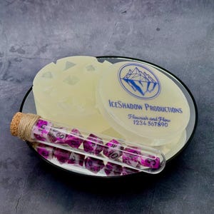 May include: A translucent off-white silicone mould for polyhedral dice with a circular lid featuring the blue text IceShadow Productions Flourish and Flow 1234567890, paired with a glass vial containing purple and white marbled dice.