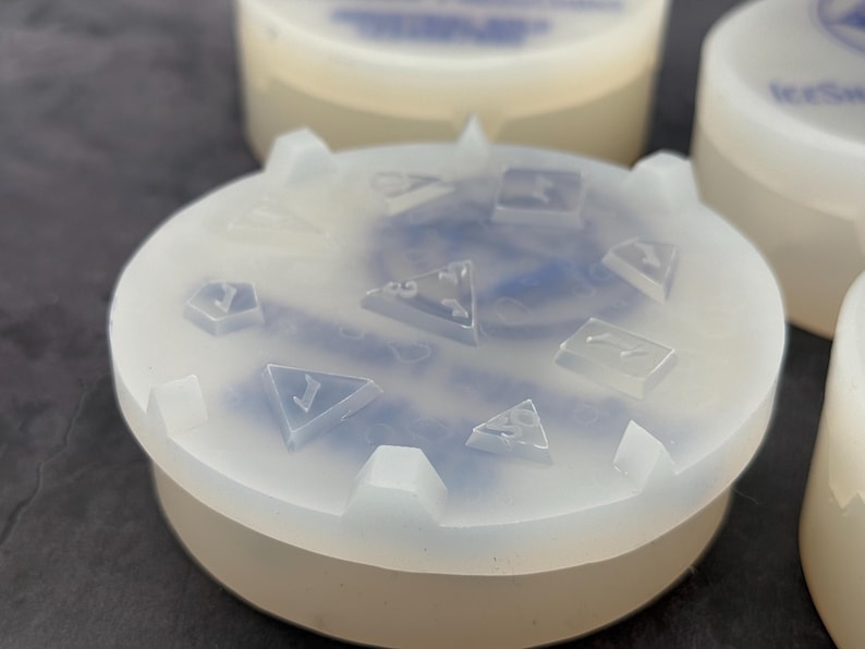 Op de afbeelding: Een transparante siliconen mal met een blauw swirl-ontwerp. De mal is rond en heeft een verhoogde rand. De mal is ontworpen om dobbelstenen te maken met verschillende nummers op elke zijde.
