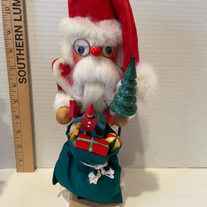 May include: A Santa Claus figurine with a red hat and coat, white beard, and glasses. He holds a green Christmas tree and a sack filled with gifts. The figurine is made of wood and fabric, and is a festive holiday decoration.