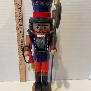 May include: A wooden nutcracker with a blue and red uniform, a black hat with gold stars, and a gold trumpet. He is holding a spear with a gold tip and a black and gold shield.