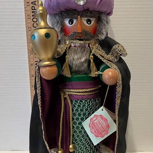 May include: A wooden nutcracker figurine of a king wearing a purple turban, green robe, and black cloak. He is holding a gold and green bottle in his right hand. The nutcracker is standing on a wooden base with a gold plaque that reads "C-M-B MELCHIOR C-M-B". The nutcracker has a label that reads "Steiff  Artifex  Original  Handcrafted  Germany".