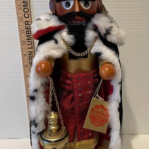 May include: A wooden nutcracker with a black beard and mustache, wearing a red and gold robe with a white fur collar and a silver and red turban with a red feather. The nutcracker is holding a gold and black oil lamp. The nutcracker is standing on a wooden base with the text "C+M+B BALTHASAR C+M+B" on it. The nutcracker has a tag that says "Steinbach Präzent" and "Handmade in Germany".