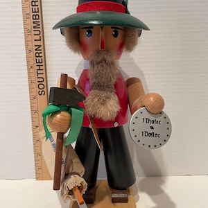 May include: A wooden nutcracker figurine of a man wearing a green hat, red shirt, and black pants. He is holding a hammer, a bag, and a broom. The nutcracker has a brown beard and mustache. The nutcracker is standing on a wooden base. The nutcracker has a silver coin attached to his belt that says "1 Thaler 1 Dollar".