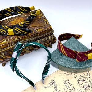 May include: Three fabric headbands with a Harry Potter theme. One is black and yellow with "Hufflepuff" text, another is green and silver, and the third is maroon and gold. The headbands are displayed on a decorative box and a stone.