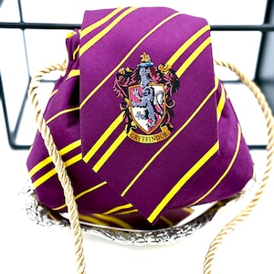 Puede incluir: Una corbata morada con rayas amarillas y un escudo de Gryffindor, colocada sobre una bolsa con cordón a juego. La corbata y la bolsa se exhiben en un plato decorativo plateado, con un detalle de cuerda dorada.