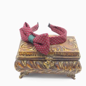 May include: A burgundy bow tie with a small white square pattern and a green center, resting on a decorative, ornate, gold-toned box. The box has a detailed, carved design and small feet.